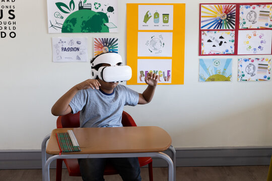 African American child wearing white VR headset sitting at classroom desk with striped pencil case - Powered by Adobe