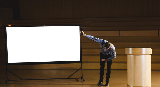 Male presenter in collared shirt bowing, holding projection screen on stage near podium, copy space