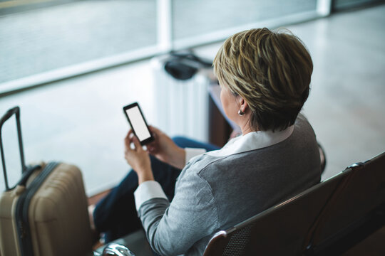 Sitting on metal bench in airport departure lounge, woman holding smartphone near spinner suitcase