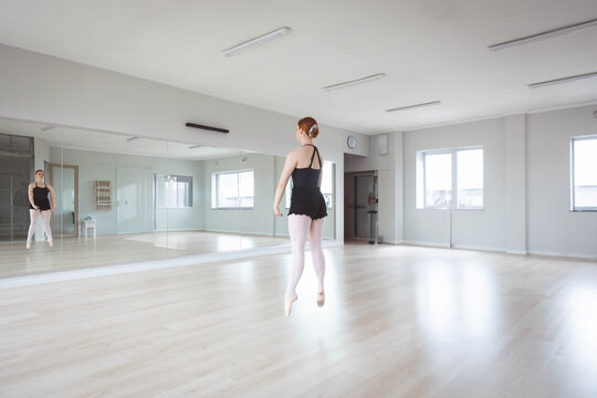 Female ballet dancer leaping in dance studio with mirror wall, barre, clock on wall, pointe shoes