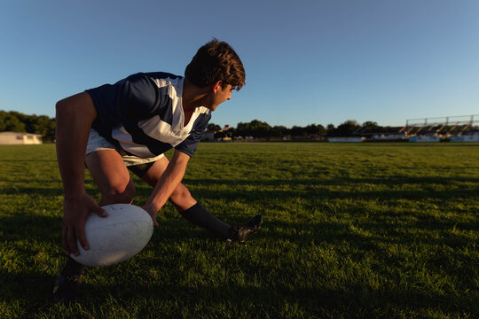 Male teenager in striped jersey crouching on field gripping rugby ball in sunlight near stands - Powered by Adobe
