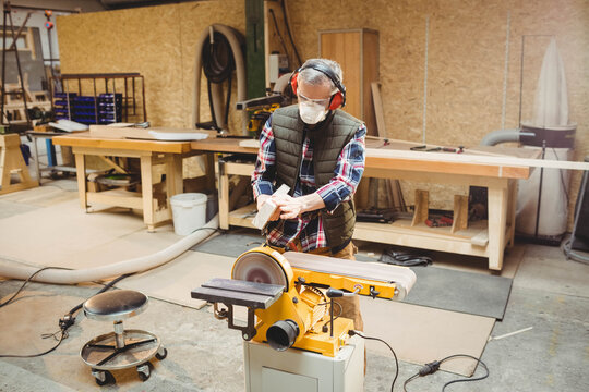 Senior man using disc sander in woodshop wearing safety gear holding wooden block near storage bins