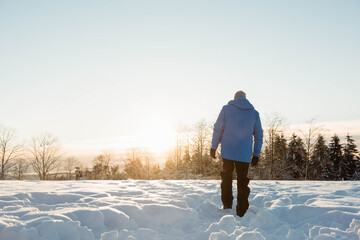 Man walking across snow-covered field at tree line wearing blue winter coat, snow pants, copy space