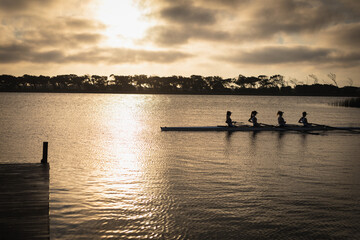 Four female rowers paddling in shell across lake at sunset beside wooden dock, copy space
