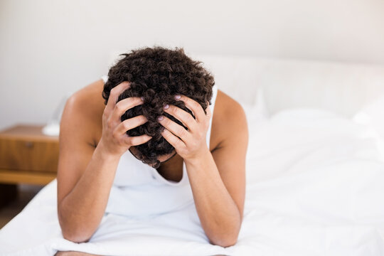 African American man sitting on white bed beside nightstand in bedroom holding head with both hands