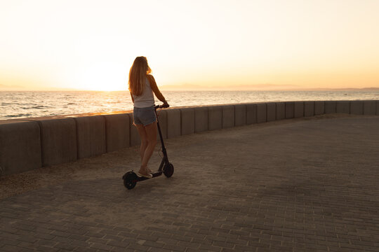 Woman standing on electric scooter facing ocean beside concrete sea wall on paved promenade
