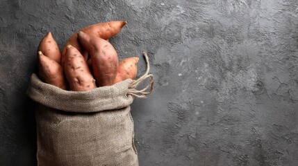 A burlap sack holds fresh sweet potatoes on a gray textured background. The sack is partially open showing the vegetables. This setup is suitable for a cooking scene or a market display.