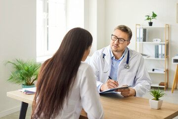 Young woman patient talking to mature male doctor in his office in clinic during medical...