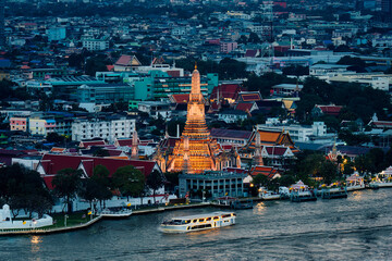 Wat Arun temple illuminated with urban houses and Chao Phraya river at dusk in Bangkok