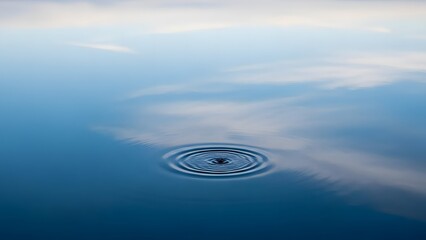 A single, perfectly circular ripple expands on the calm, reflective surface of deep blue water, mirroring the soft, cloudy sky above.