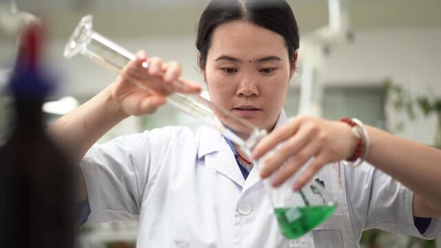 Female researcher carefully pouring green liquid from a graduated cylinder into a beaker in a laboratory setting