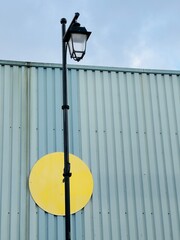 Street lamp stands next to a yellow circle on a building during daylight under a cloudy sky