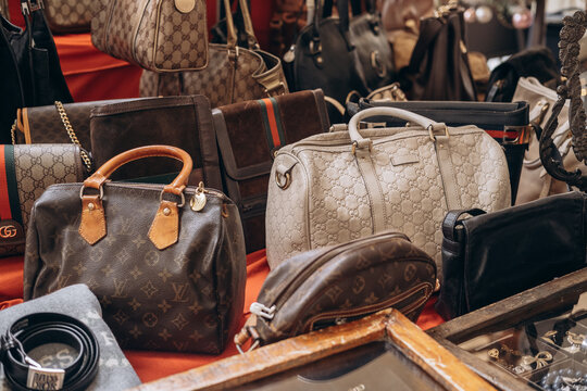 A vibrant display of luxury handbags from Gucci and Louis Vuitton on a bustling market stall. Leather textures, monogram patterns, vintage market. Milan Italy January 3, 2026 