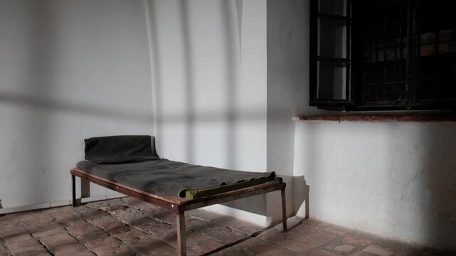 An old prison bed in an authentic prison cell. the shadows of the prison door are casting on the floor and walls as the door opens. a 4K video clip.