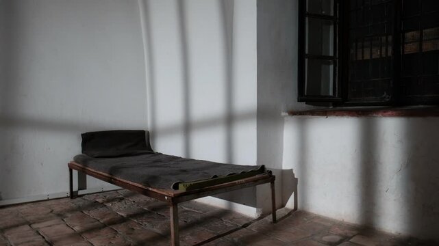 An old prison bed in an authentic prison cell. the shadows of the prison door are casting on the floor and walls as the door closes and back opened. a 4K video clip.