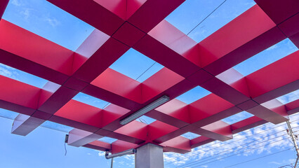 Red geometric grid canopy structure against a blue sky. Modern architectural pattern with steel beams for outdoor pavilion, minimalist design, and abstract background.