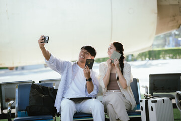 Naklejka premium Young Asian couple taking a selfie while holding passports at an airport terminal, representing travel, tourism, international journey, and happy vacation preparation.