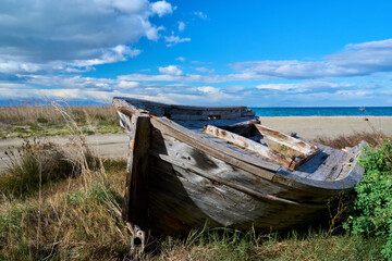 Abandoned wooden fishing boat on a sandy beach with dry grass, blue sea and dramatic cloudy sky. Coastal landscape, weathered wood texture, seaside atmosphere.