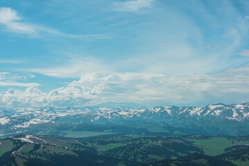 Aerial sunlit top view above forest hills and big snowy mountain range under lush clouds in changeable weather. Scenic hilly alpine landscape with sunlight and shadows. Partly cloudy in high mountains