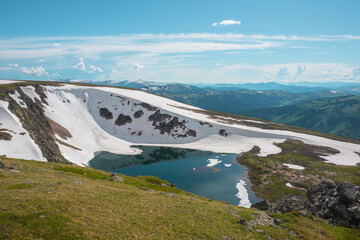 Scenic sunlit landscape with alpine lake in rocky snowy cirque near stone hill top during thaw. Ice floats in mountain lake among rocks and snows with view to forest mountain range under cloudy sky. © Daniil