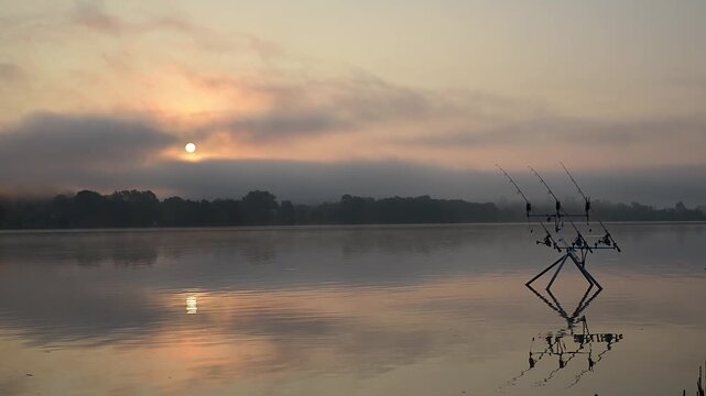 Fishing adventures, carp fishing on lake with the sun peeking through the fog and clouds. Rod pod with fishing rods and bite alarms at sunrise with mist. Quiet and relaxing atmosphere in nature