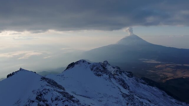 Aerial view of the snow-covered Iztaccihuatl volcano at dawn with Popocatepetl in the background emitting smoke in Mexico - 4K Video