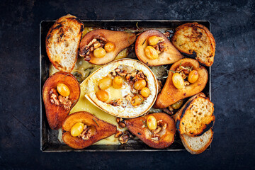 Oven baked camembert cheese with caramelized glazed pears, walnuts and grapes and baguette served as top view on rustic metal sheet