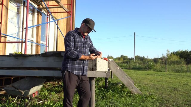 A grown man stands near a wooden house. Construction. Dressed in a shirt and a cap. With glasses and a beard. Holding a smartphone. Business. Farmer. Summer day. Sunset.