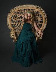 Obraz premium Portrait of beautiful blonde female model wearing royal green ballgown, fantasy crown. Sitting pose in throne chair isolated on dark studio background.