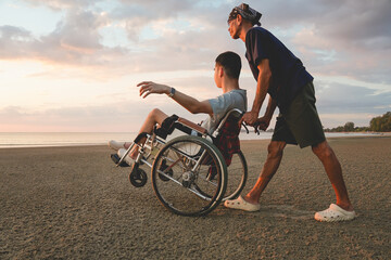 Asian elderly caregiver pushing wheelchair with disabled family member at the beach during sunset. Loving family support, inclusion, accessibility, and emotional outdoor moment.