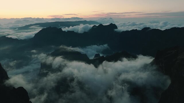 Aerial drone shot of rugged rocky silhouettes rising above a sea of clouds in Madeira during late sunset. The landscape is mostly in cool, dark tones, while the sky above horizon retains warm colors