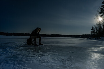 A dramatic night photograph of an ice fisherman sitting on a frozen lake under a bright moon. The scene is captured in Finland.