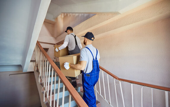 Movers carry boxes up stairs during relocation. Uniformed workers climb a stairwell with cartons, ensuring safe handling and logistics delivery. Efficient moving service, transport and teamwork.