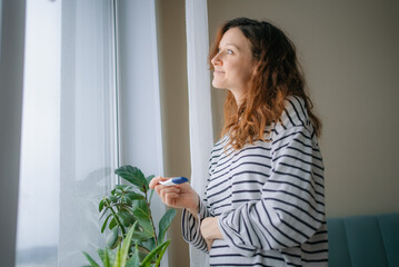 Woman holding pregnancy test, looking hopeful and smiling, anticipating results with excitement, contemplating new life and family planning future next to a window