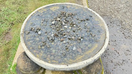 Fototapeta premium A circular stone basin filled with clear water and a pile of natural pebbles. Situated in an open garden area, it faces a backdrop of green grass and a dirt path.