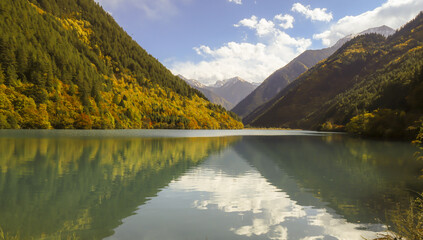 Serene mountain lake reflects surrounding lush green and autumn colored forests under partly cloudy sky. Snow capped peaks are visible in distance, enhancing tranquil atmosphere