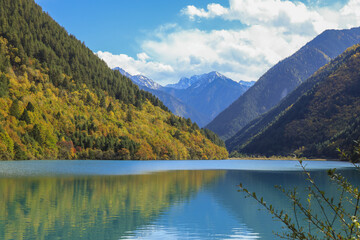 Fototapeta premium Serene mountain lake surrounded by lush green forests and distant snow capped peaks under bright blue sky. calm water reflects vibrant landscape, creating peaceful atmosphere