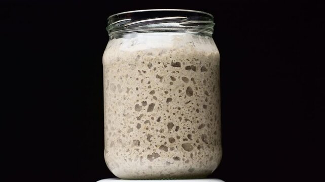 Sourdough starter in a glass jar rotating on a black background. Active liquid yeast fermentation process with bubbles.