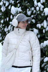 A woman in a white puffer jacket, grey cap, and white earmuffs stands before a snow-dusted evergreen bush, looking off-camera with a gentle smile during a bright winter day. © Anastasiia