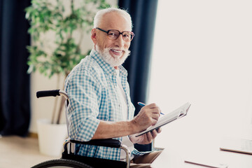 Older man in wheelchair writes in a notebook at home; a warm, inclusive moment of everyday life and creativity in a bright living room