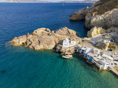 Aerial view of the whitewashed houses and boat garages nestled against the rocky coastline with turquoise waters, Firopotamos, Milos, Greece.