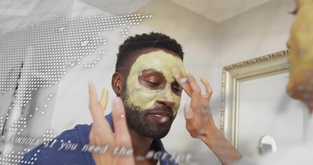 Standing adult man in blue robe getting facial mask smoothed by partner at home vanity, mirror