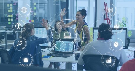 High-fiving standing woman in sleeveless top leaning over shared desk at open office, with laptops