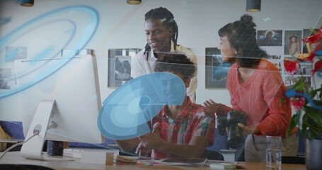 Collaborating three colleagues pointing at desktop in creative office, coral sweater, DSLR camera