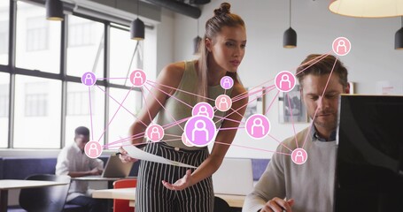 Leaning woman in sleeveless top holding docs, gesturing at monitor in office, pink network overlay