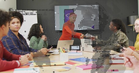 Shaking hands presenter in orange top with colleague in plaid at conference table, laptops visible