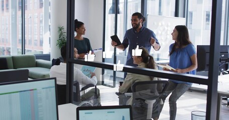 Presenting man in dark shirt leading meeting in glass room, with tablet, charts and monitors