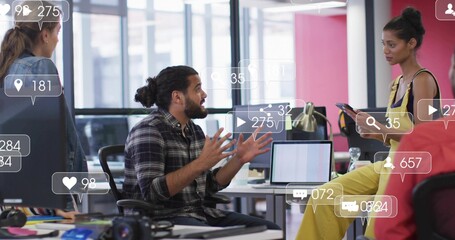 Gesturing man in plaid shirt speaking in open office, woman in yellow holding phone, social icons