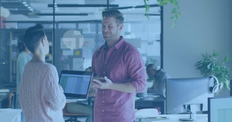 Talking mid adult man in pink buttondown shirt holding smartphone at open office, with open laptop
