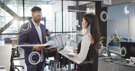 Discussing two colleagues wearing blazers exchanging laptop and documents in open-plan office, HUD
